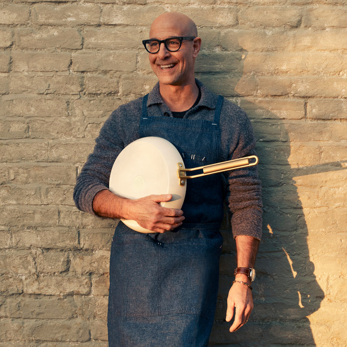 Man in glasses and apron holds a Stanley Tucci™ ceramic nonstick pan, standing by a brick wall.
