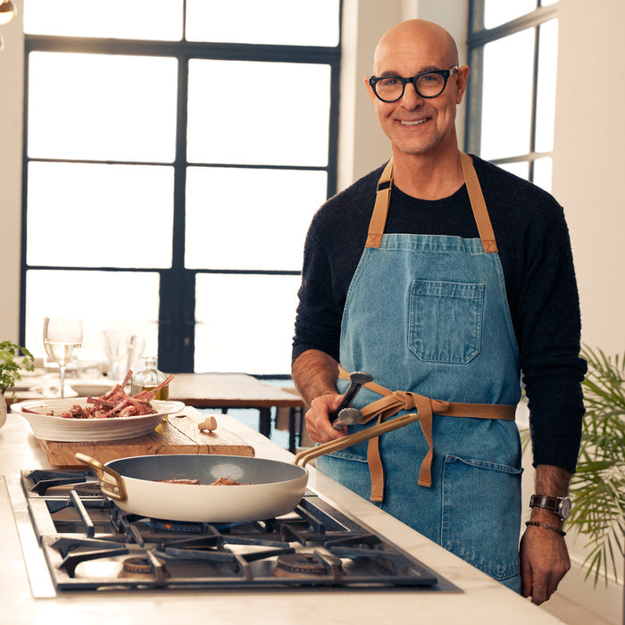 Man in glasses and denim apron using Stanley Tucci 11-piece cookware set in a modern kitchen.