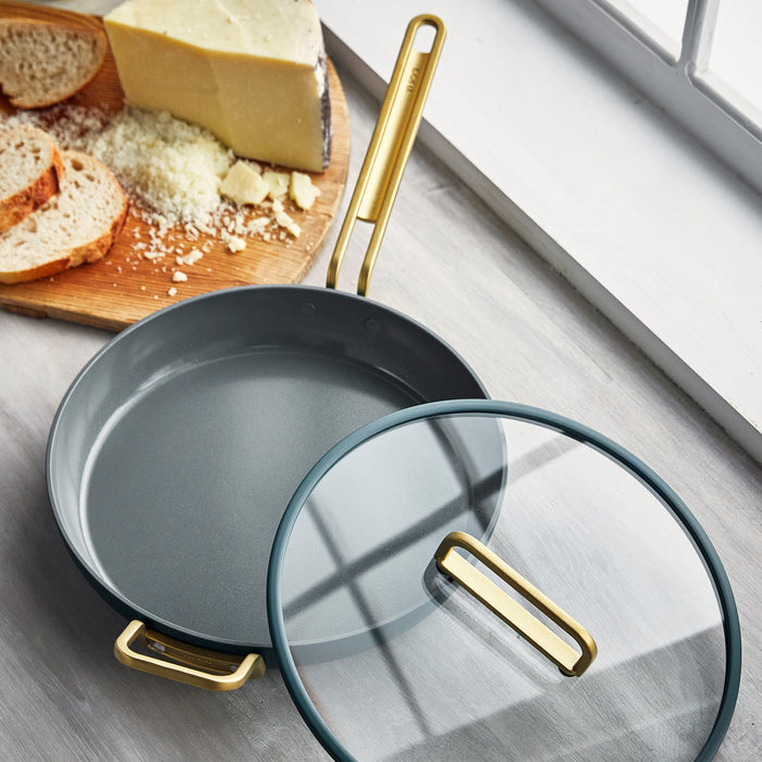 Venetian teal ceramic nonstick frypan with lid, next to sliced bread and cheese on a wooden cutting board.