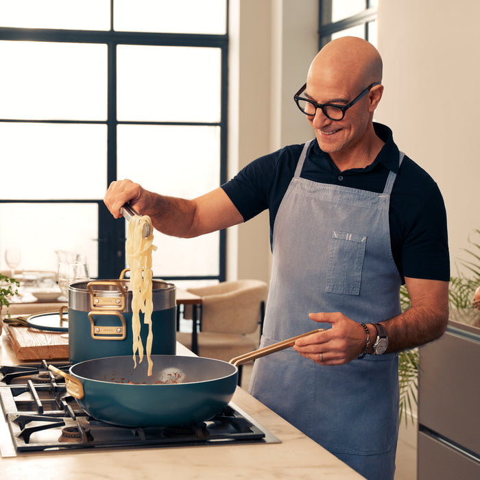 Man in glasses and apron lifts pasta using Stanley Tucci ceramic nonstick cookware set with multiple pieces.