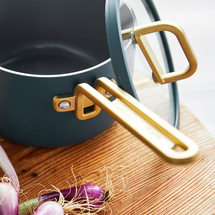Venetian teal saucepan with gold handle and lid next to fresh shallots on a kitchen surface.