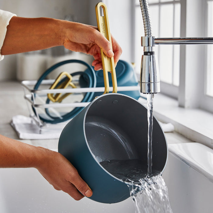 Venetian Teal ceramic saucepan being rinsed under water, with other clean pots drying nearby on the counter.