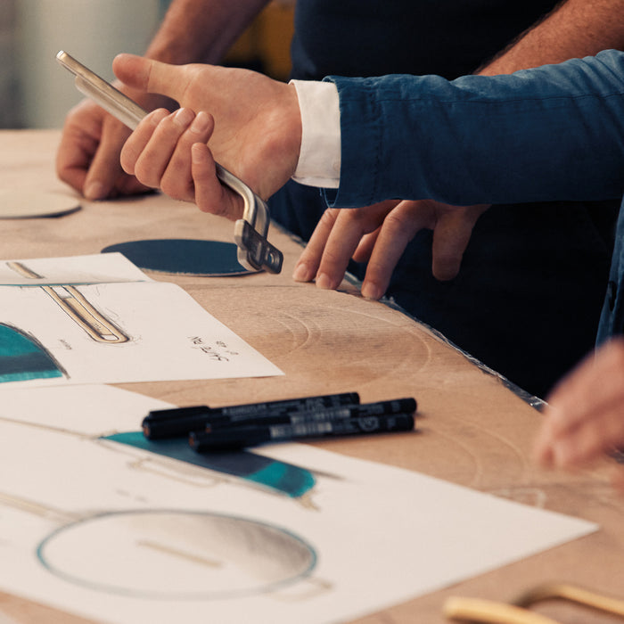 Hands holding a phone above technical drawings with a Venetian Teal saucepan visible.
