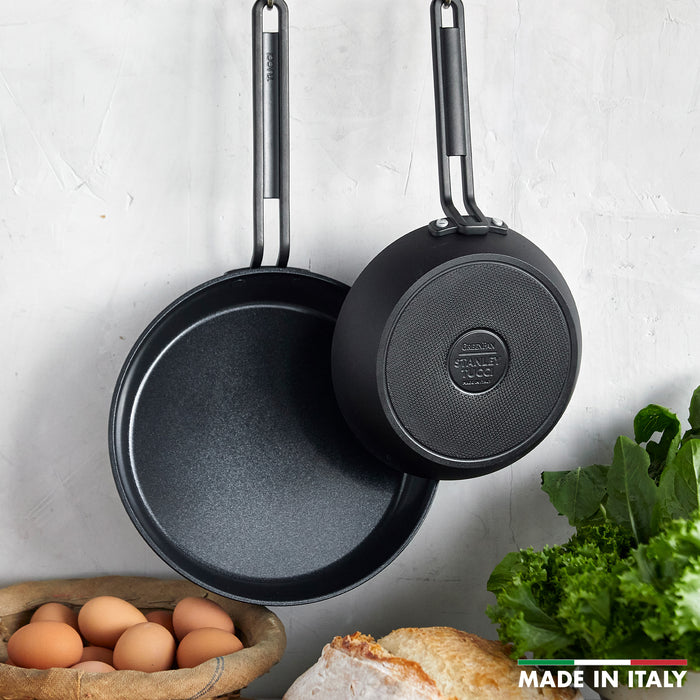 Two black ceramic nonstick frypans, 8 and 10, hanging above eggs, bread, and greens on a counter.