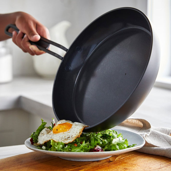 Hand tilting a white ceramic frypan to slide a sunny-side-up egg onto a bowl of salad.