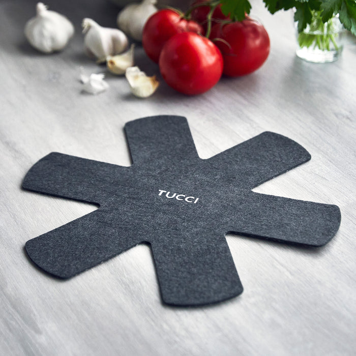 Black felt pan protector with TUCCI text next to tomatoes, garlic, fresh herbs, and a ceramic nonstick frypan set.
