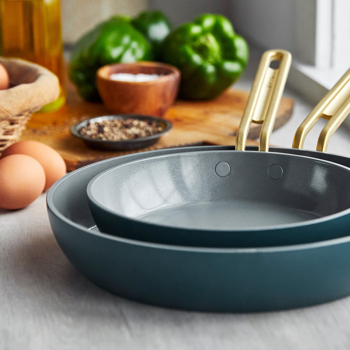 The Stanley Tucci™ Ceramic Nonstick 8" and 10" Frypan Set in Venetian Teal, featuring gold handles, is stacked on a kitchen counter with eggs, green bell peppers, oil, and a wooden seasoning bowl nearby. PFAS-free for safer cooking.