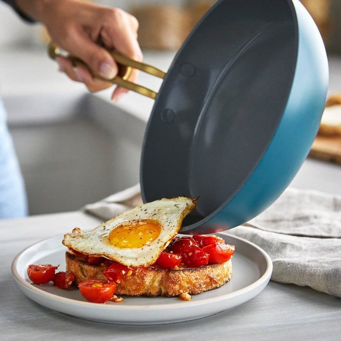 Hand sliding a fried egg from a teal ceramic nonstick 8-inch frypan onto toast.