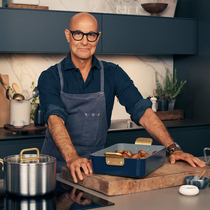 Man in glasses and apron holding food in a blue Stanley Tucci lasagna pan in a modern kitchen.