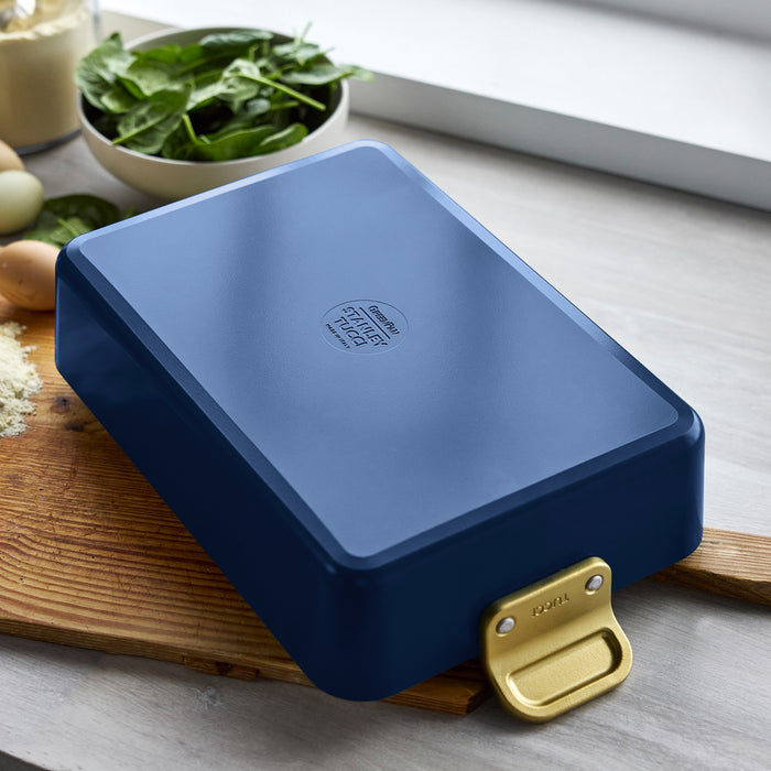 Marino blue lasagna pan with gold handle on a wooden board next to fresh vegetables and herbs.