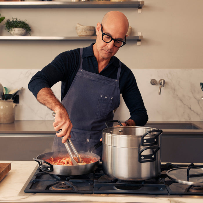 Man wearing glasses and apron cooking pasta in a Stanley Tucci 12-inch nonstick frypan with lid on stove.