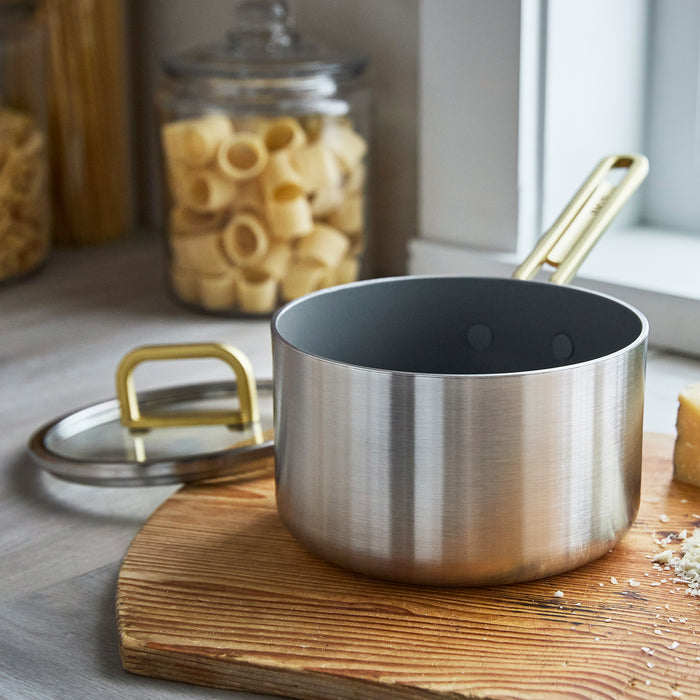 Stainless steel saucepan with champagne handles and lid beside pasta and cheese on a counter.