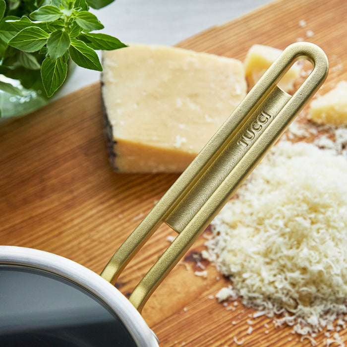 2-quart stainless steel saucepan with champagne handles on a wooden board, next to grated cheese and fresh basil leaves.