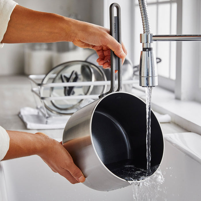 A stainless steel saucepan with lid is being rinsed under running water at a kitchen sink.