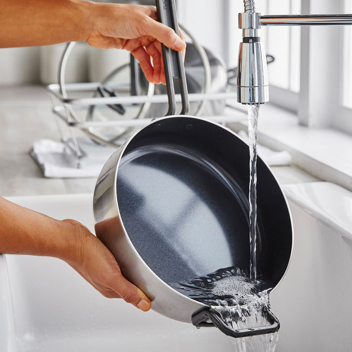 A stainless steel sauté pan with ceramic coating and lid being rinsed under running water in a kitchen sink.