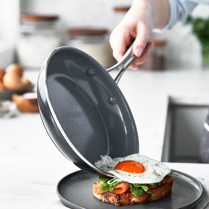 Hand sliding fried egg from ceramic nonstick pan onto toast topped with greens, sliced tomatoes, and crispy shrimp.