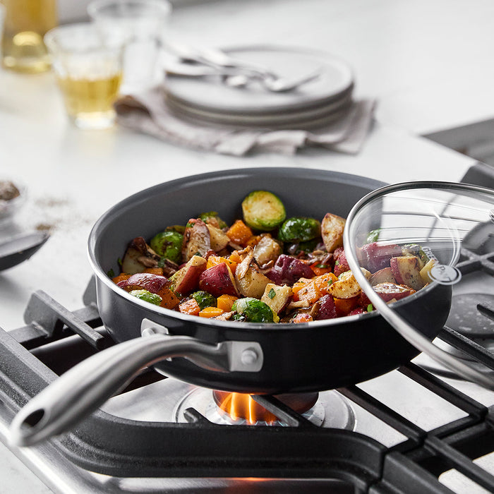 Ceramic nonstick pan with veggies and meat cooking on a gas stove, glass lid partially covering the pan.