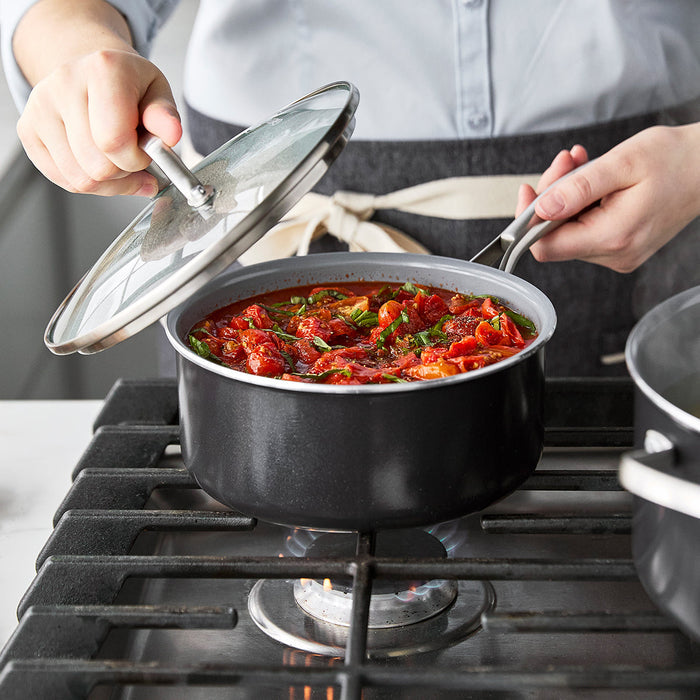 Hand lifting lid from white ceramic nonstick pot with tomato stew and seared meat simmering on a stovetop.