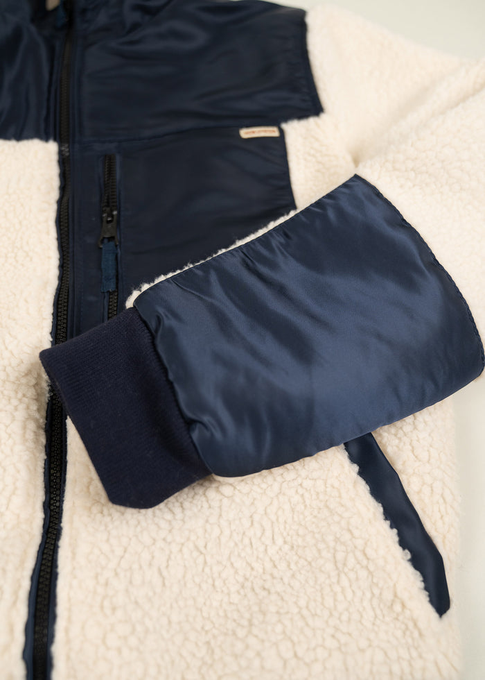 Close-up of a cream sherpa jacket with navy nylon accents, featuring a zipper and pocket, showcasing texture and design details.