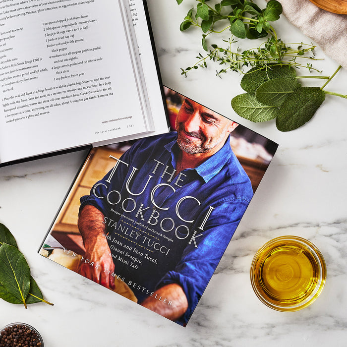 Tucci ceramic frypan set with two pans displayed on a table beside herbs, olive oil, and an open cookbook.