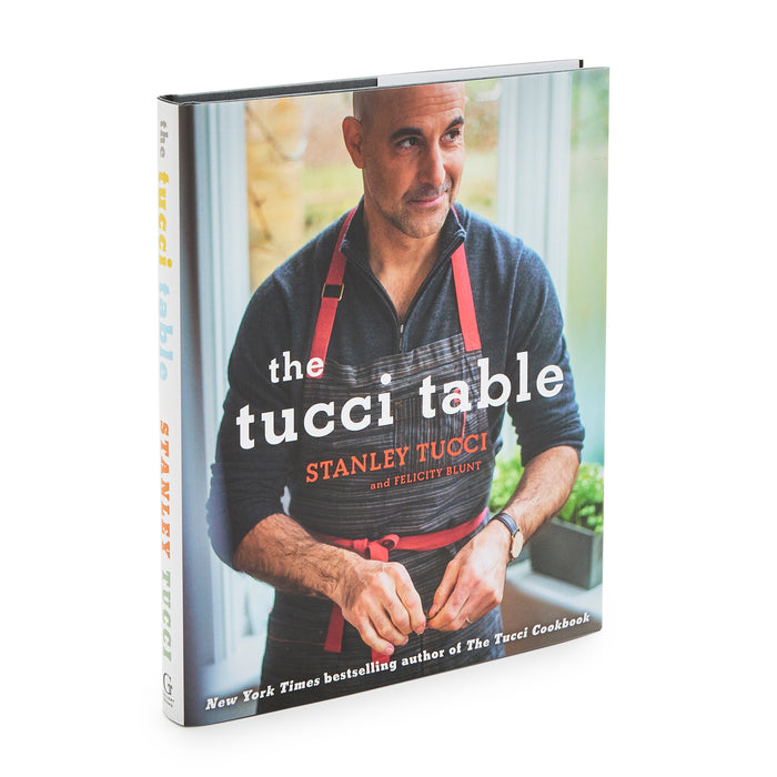 Stanley Tucci wearing an apron stands by a window holding The Tucci Table Cookbook.