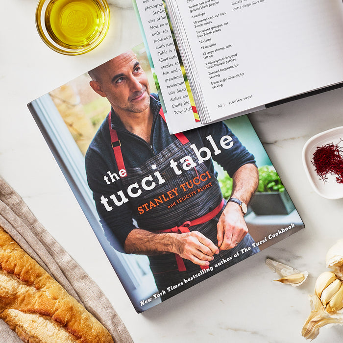 Open cookbook with sliced bread, olive oil in a bowl, garlic cloves, and saffron threads on a light surface.