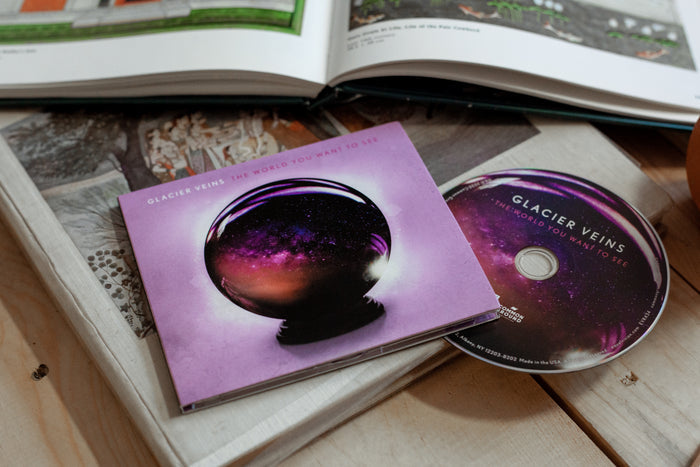 Purple square CD with white font that says GLACIER VEINS. Next to that there is more font that says THE WORLD YOU WANT TO SEE in white. Below that there is a purple crystal ball. Peeking out of the cover is a CD with the design of the crystal ball. Both are laying on top of a wooden surface.