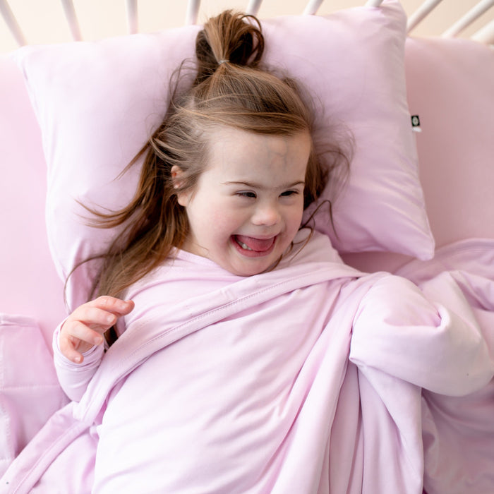 Young smiling girl laying in bed underneath a Toddler Blanket in Thistle 1.0 with her head resting on a toddler pillow with matching pillowcase