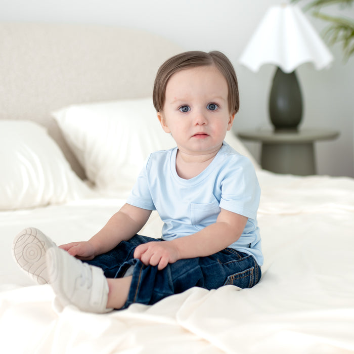 Young toddler sitting on a white bed wearing the Toddler Crew Neck Tee in Breeze with dark wash jeans