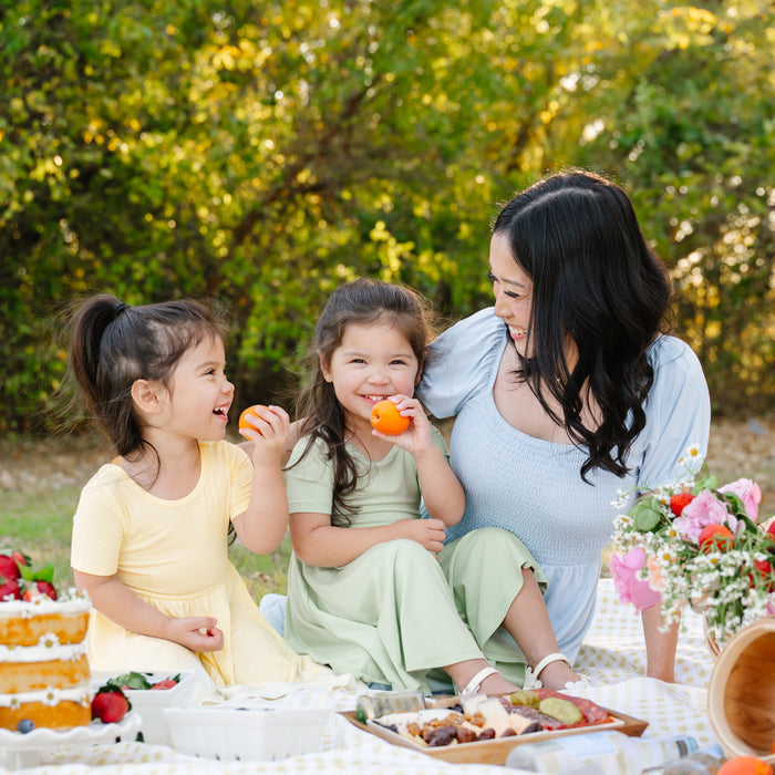 Two girls wearing the Twirl Dress in Basil and Chamomile sitting having a picnic with their mother wearing the Women's Smocked Dress in Breeze