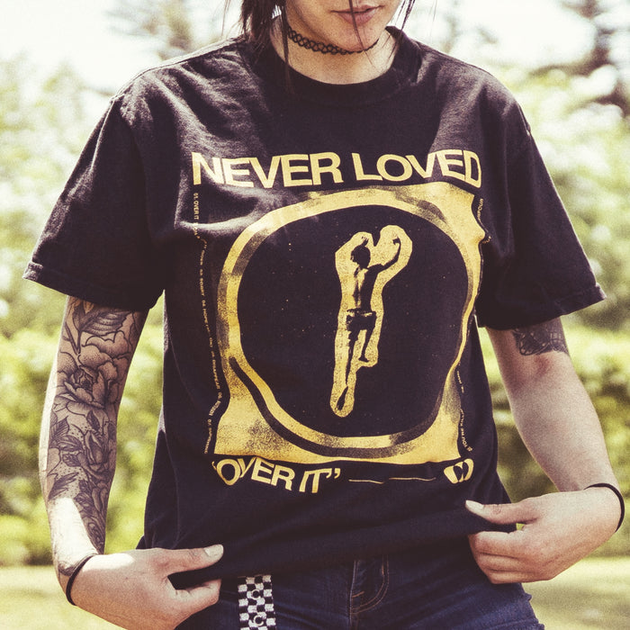 Black short sleeve shirt with yellow text that says NEVER LOVED across the chest. Below that there is a yellow drawing of the outline of a boy jumping. Below the drawing there is more yellow text that says OVER IT. An individual is modeling the shirt and standing on grass. There are trees in the background.