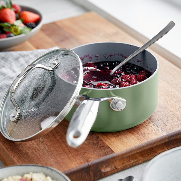 Sage green ceramic saucepan set with lids, filled with berry compote, on a wooden board beside fresh fruit.