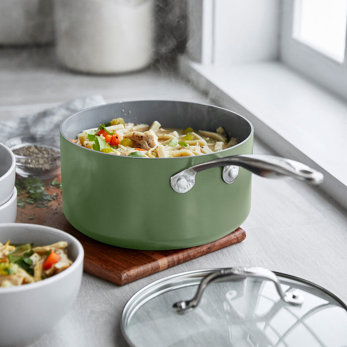 Sage green saucepan set with lids on a wooden trivet near a window, cooking pasta and vegetables.