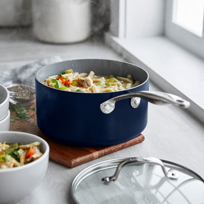 Ceramic nonstick saucepan set with lids, gray, two pieces, on a wooden board by a window, filled with pasta and veggies.