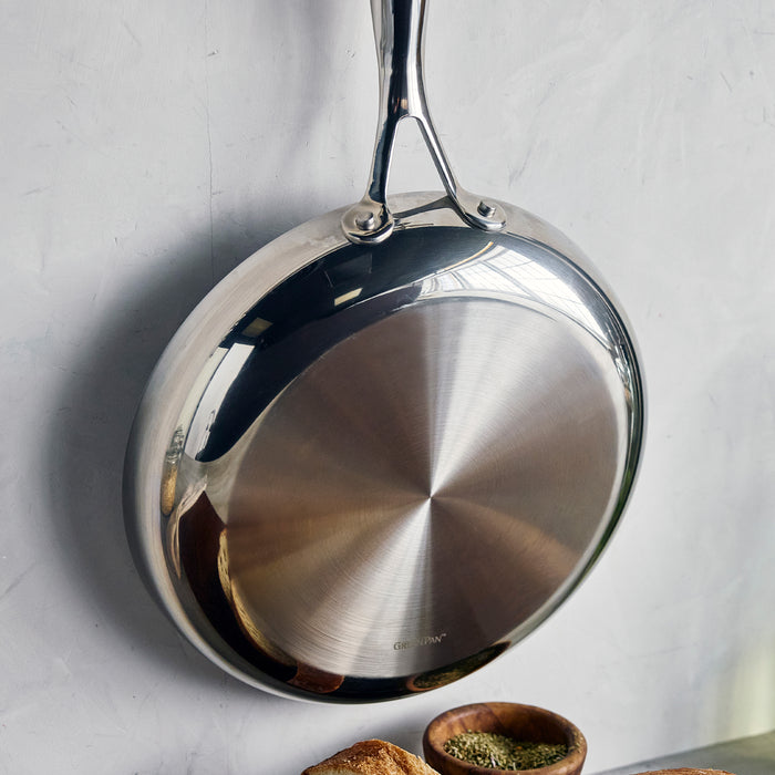 Stainless steel frypan hanging on a light wall above a small bowl of herbs and several bread slices.