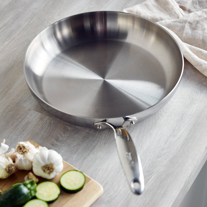 Stainless steel 12-inch frypan next to sliced zucchini and garlic on a wooden cutting board.