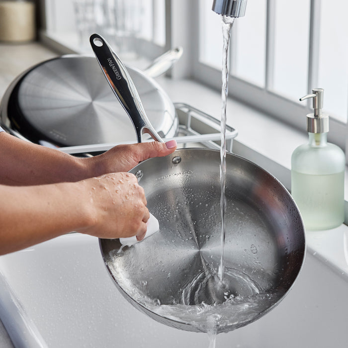 Washing a stainless steel 12-inch frypan under running water next to a kitchen sink soap dispenser.
