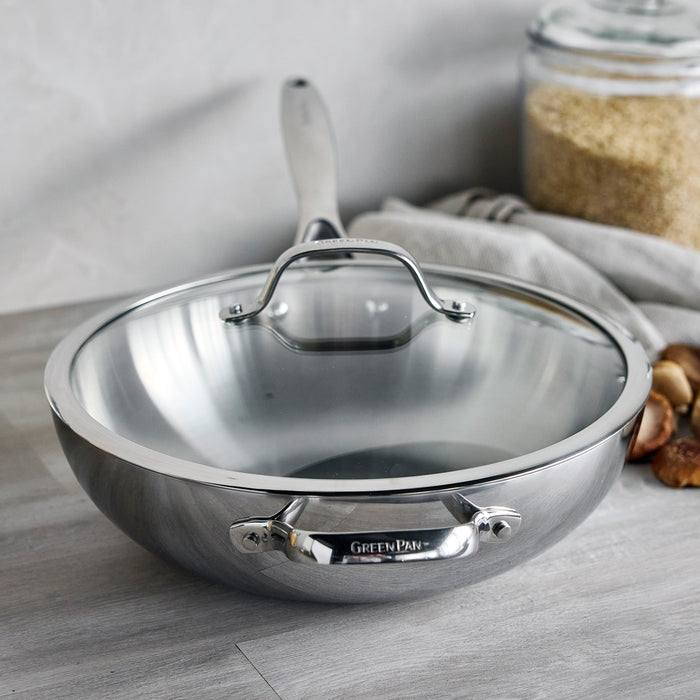 Stainless steel 12-inch wok with lid on a counter, next to a jar of grains and garlic.