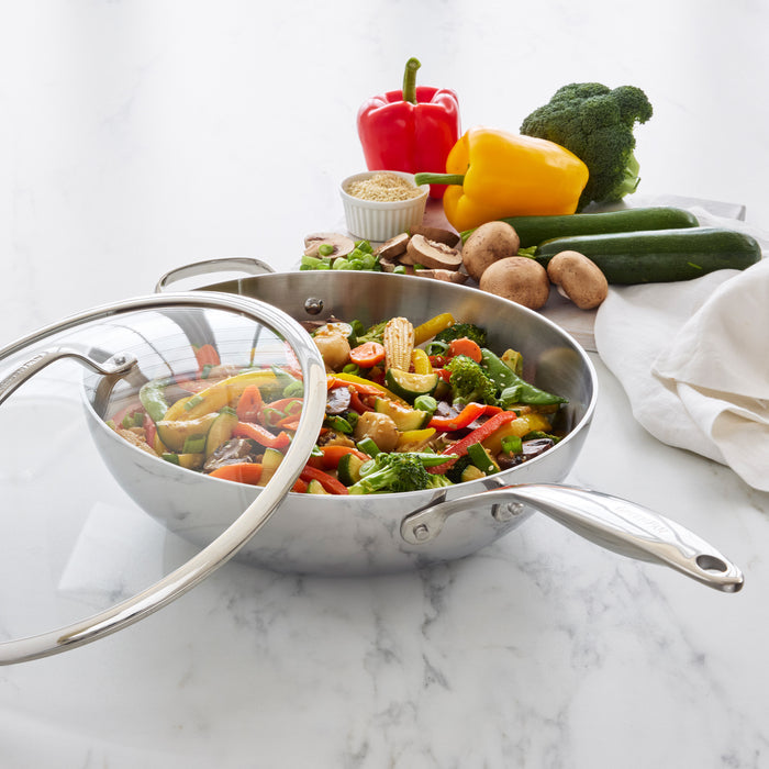 Stainless steel 12-inch wok with lid, filled with vegetable stir-fry, on a marble countertop.