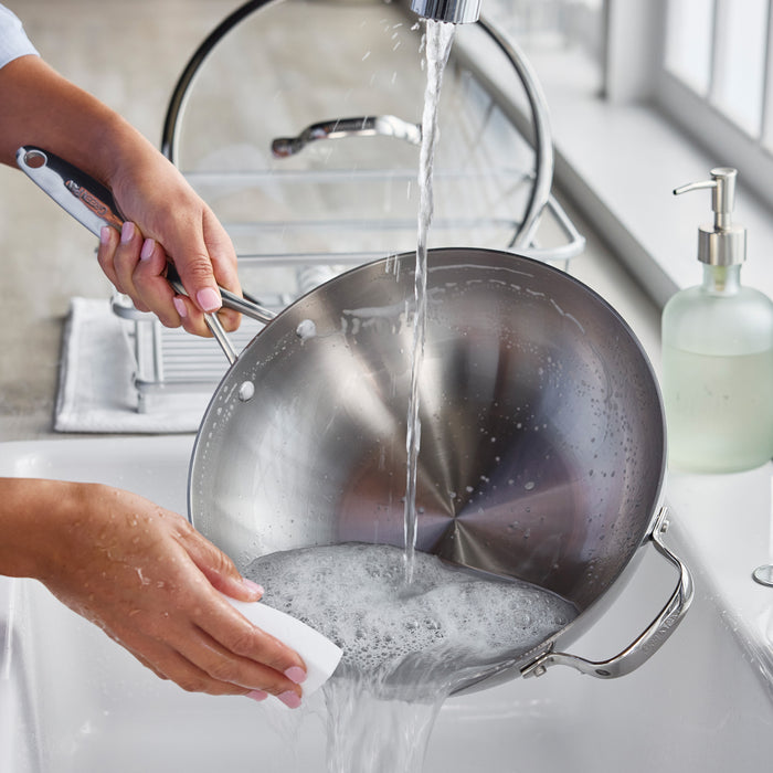 Washing a soapy stainless steel wok with lid at a kitchen sink using a white sponge.