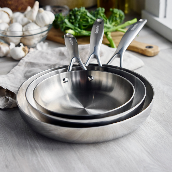 Three stainless steel frypans 8, 9.5, 11 nested on a counter with assorted vegetables and a kitchen towel nearby.