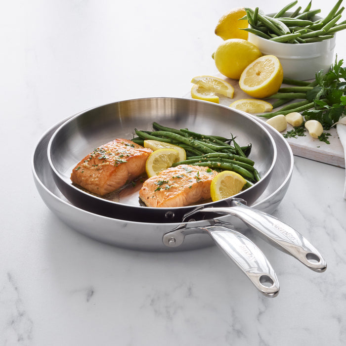 Salmon fillets and green beans in a stainless steel frypan, with lemons and assorted vegetables on the counter nearby.