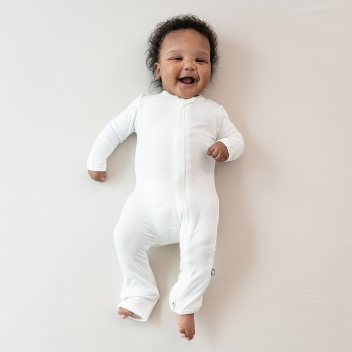 Smiling baby laying on Oat surface wearing Kyte Baby Zippered Rompers Zippered Romper in Cloud